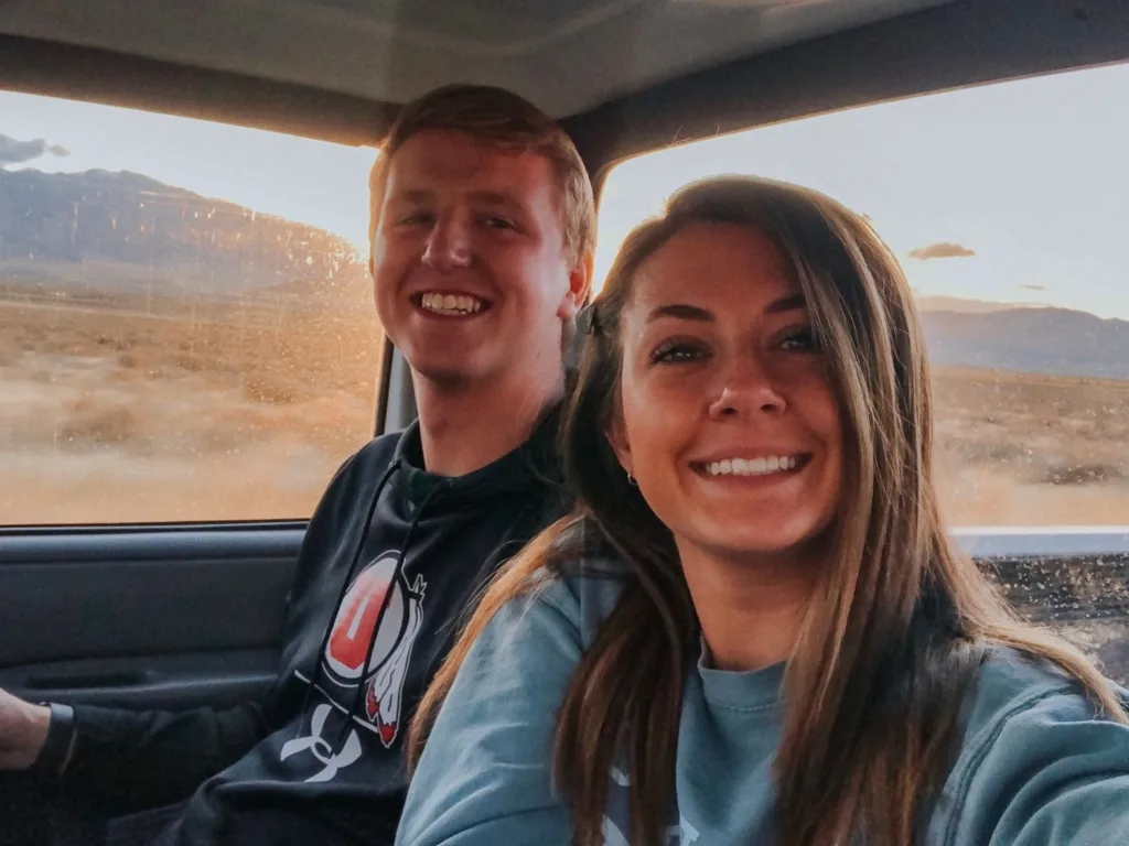 Couple smiling for a selfie inside a pickup truck with a desert landscape visible through the windows.