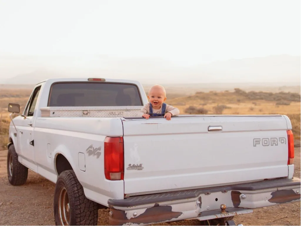 Baby smiling while leaning over the tailgate of a white Ford pickup truck in an open desert area.
