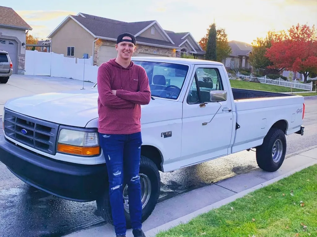 Man standing with arms crossed beside a white Ford F-150 parked on a residential street.