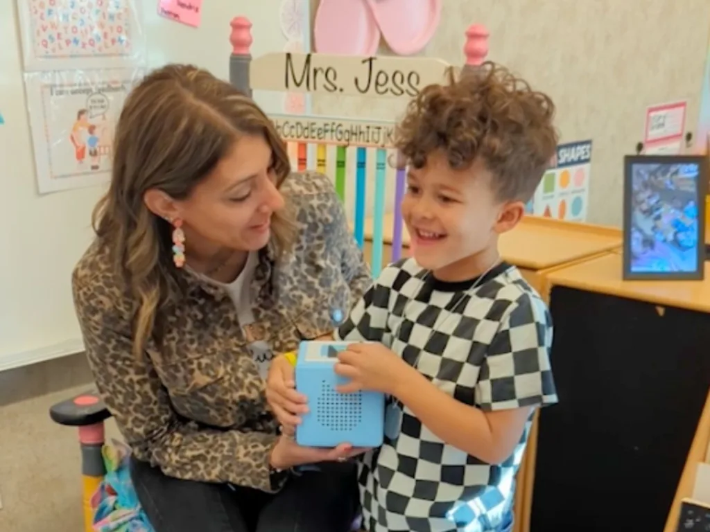 A preschool teacher holds a blue Toniebox while a young boy in a checkered shirt smiles beside her in a classroom.