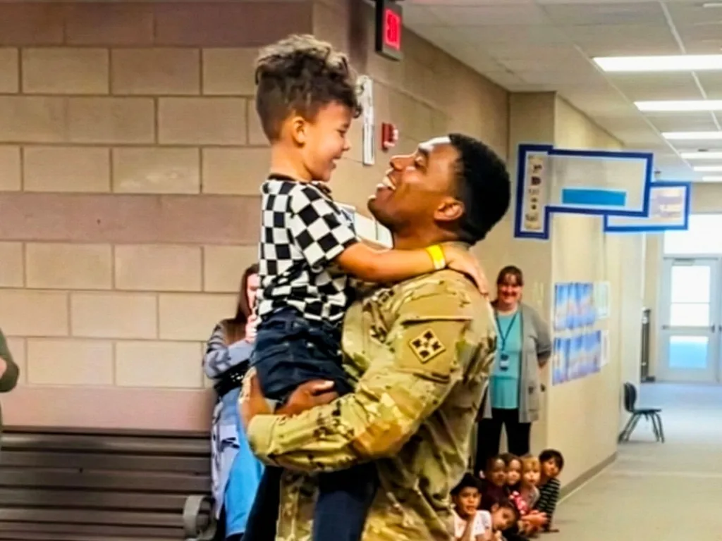 A soldier in uniform lifts a young boy in a checkered shirt for a hug in a school hallway as children watch nearby.