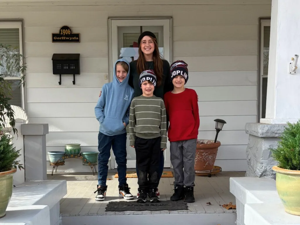 A woman stands on a front .porch with three boys in front of a house