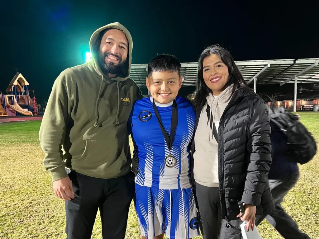 A boy in a blue soccer uniform and medal smiles while standing between two adults on a lit sports field at night.