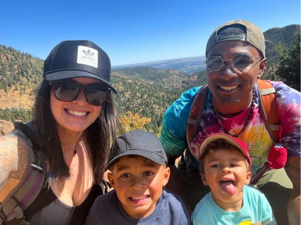 A family takes a sunny outdoor selfie during a hike with mountains and a valley in the background.