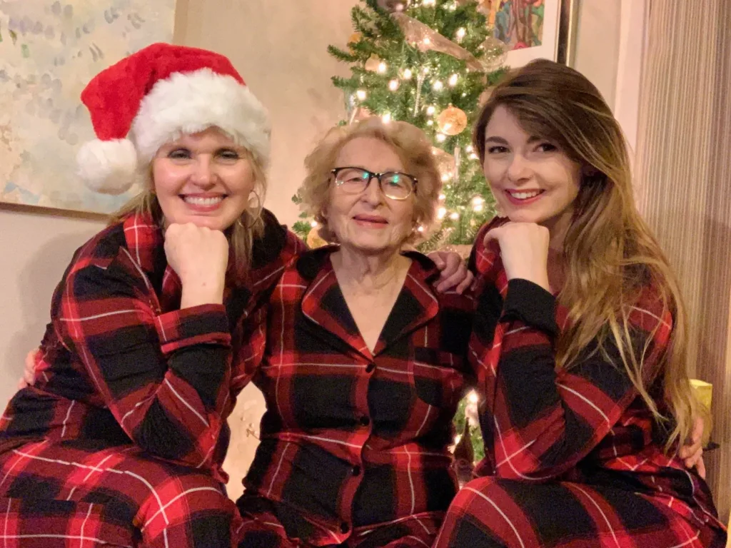 Three women in matching red plaid pajamas pose smiling in front of a lit Christmas tree.