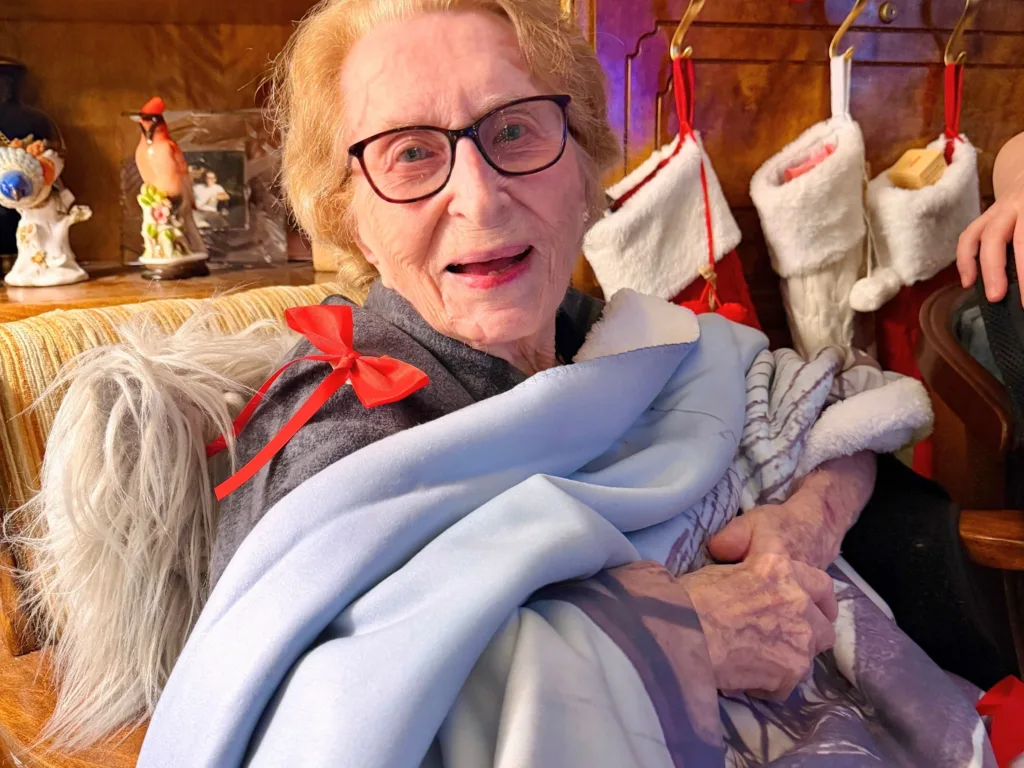 Smiling elderly woman wearing glasses holds a photo blanket while a small dog with a red bow sits beside her.