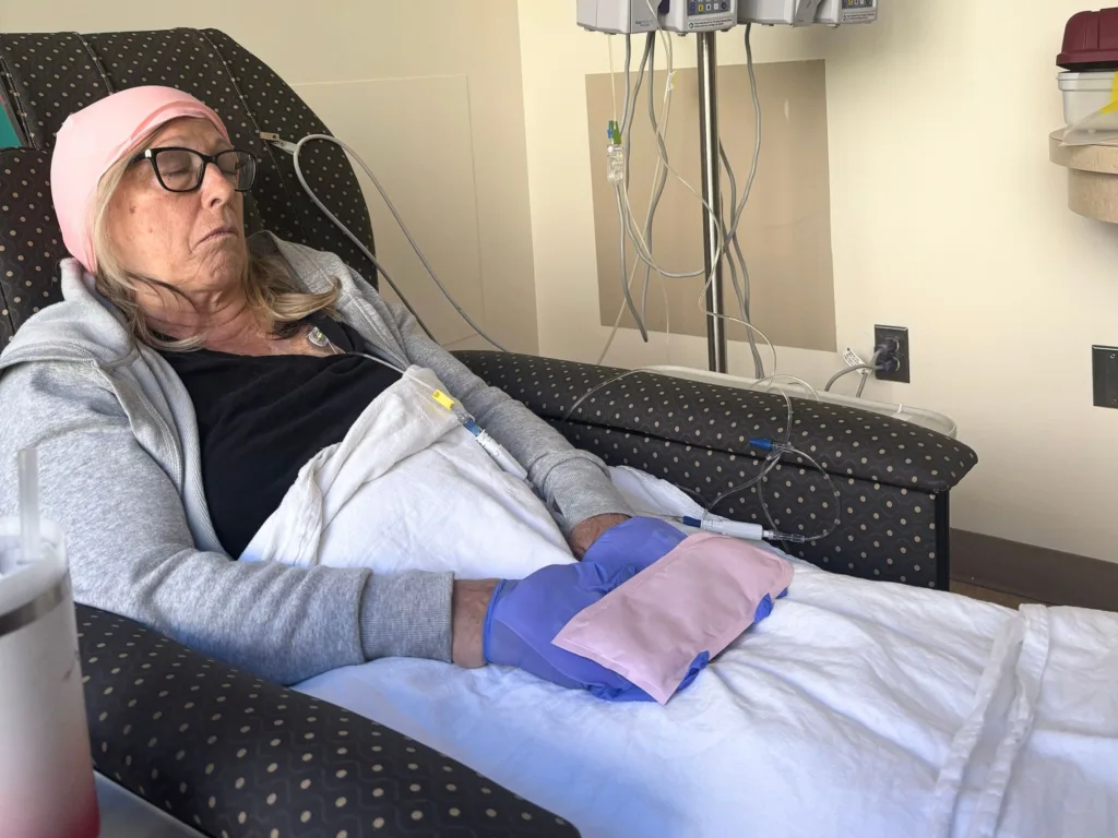 Teresa rests in a medical recliner wearing a pink cap and gloves during cancer treatment.