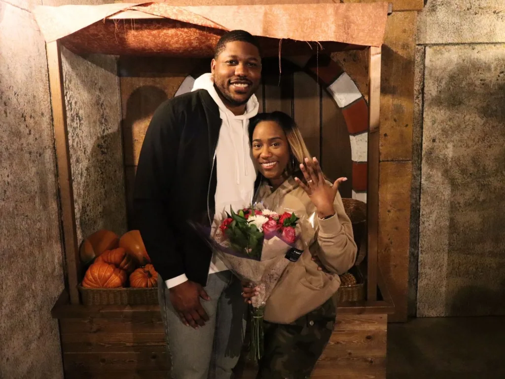 Jazmine smiles and holds up her hand to show her engagement ring while standing with Michael and a bouquet of flowers.