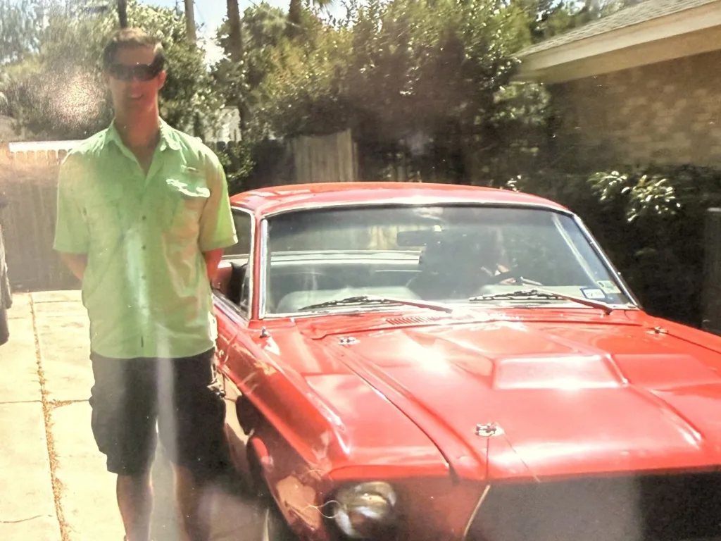 Kyle stands beside a red classic Ford Mustang in a driveway on a sunny day.