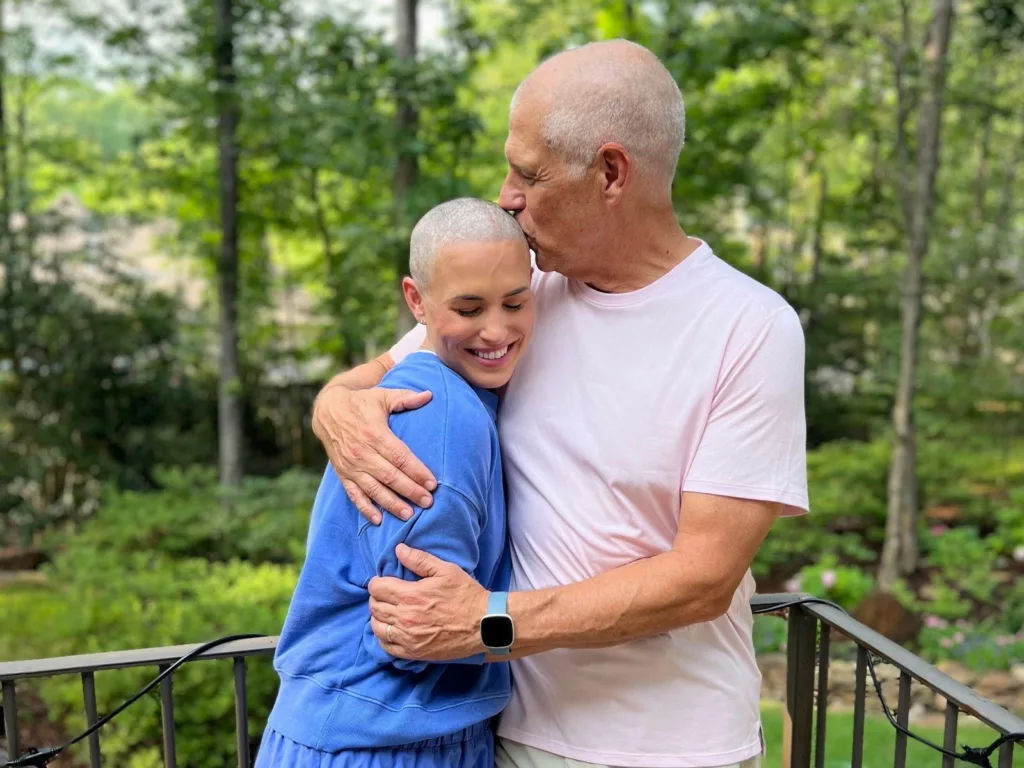 Rob hugs Alice outdoors and kisses the top of her head as she smiles, with trees and greenery in the background