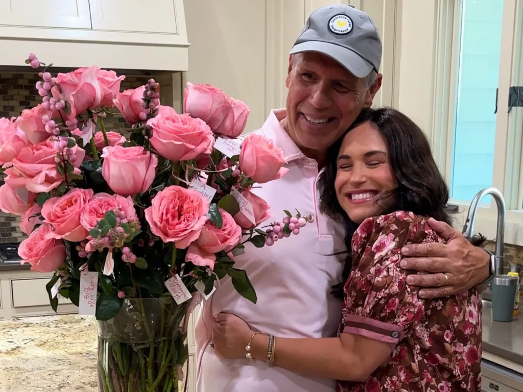 Rob hugs Alice in a kitchen beside a large vase overflowing with pink roses, with small handwritten notes attached to stems