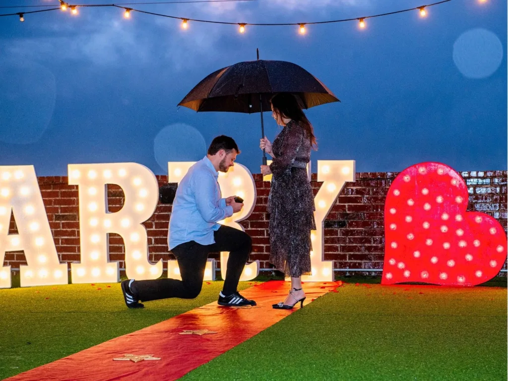 Hunter kneels with a ring as Kim stands under an umbrella near a glowing “Marry” sign.