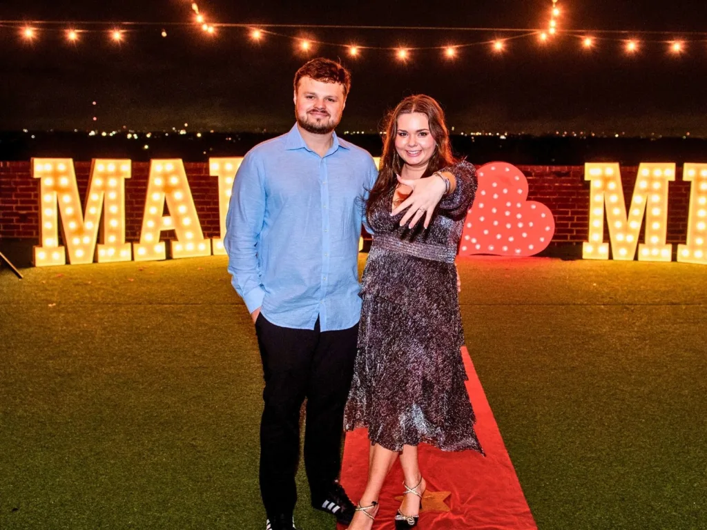 Hunter and Kim pose in front of a lit “Marry Me” display as Kim holds up her ring hand.