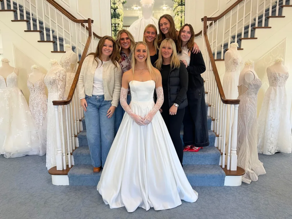 Sitori stands on stairs in a wedding dress with friends inside a bridal shop surrounded by mannequins in gowns.