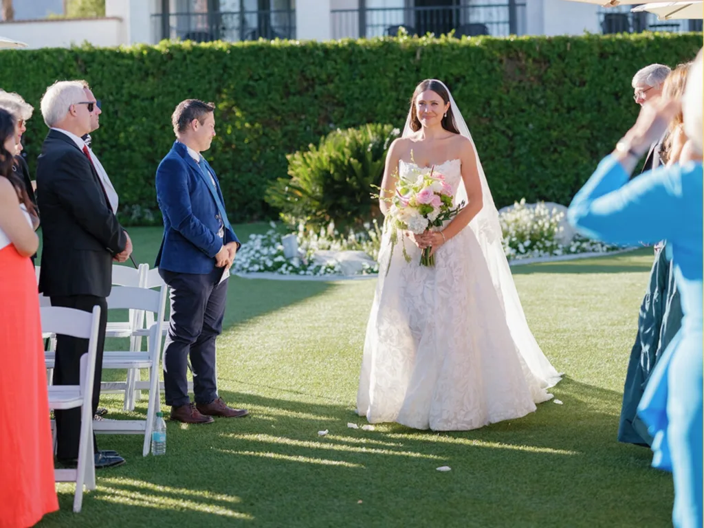 Lizzie Dean stands facing the front of an outdoor wedding ceremony in a strapless gown and veil, holding a bouquet.