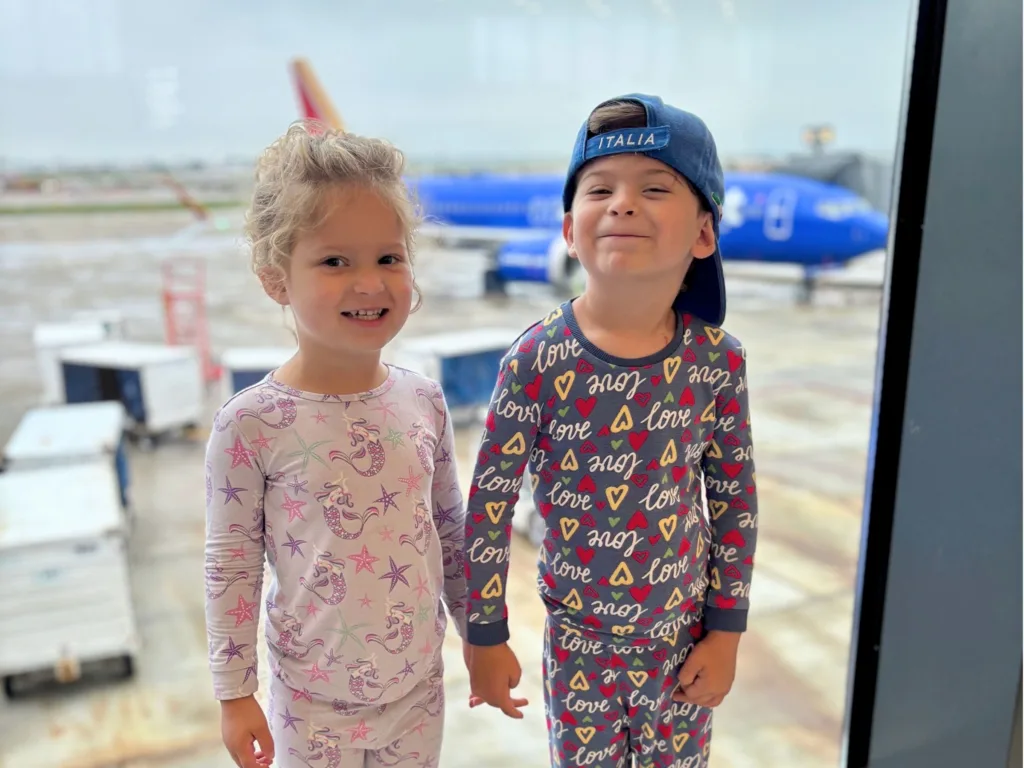 River and David stand smiling by an airport window in matching pajamas before boarding a flight.