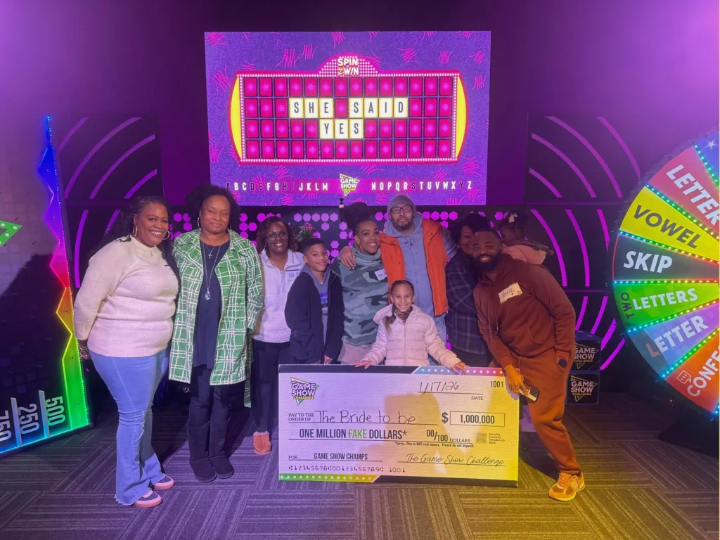 Alicia and Andre pose with their family and friends in front of a game board that reads “She Said Yes.”