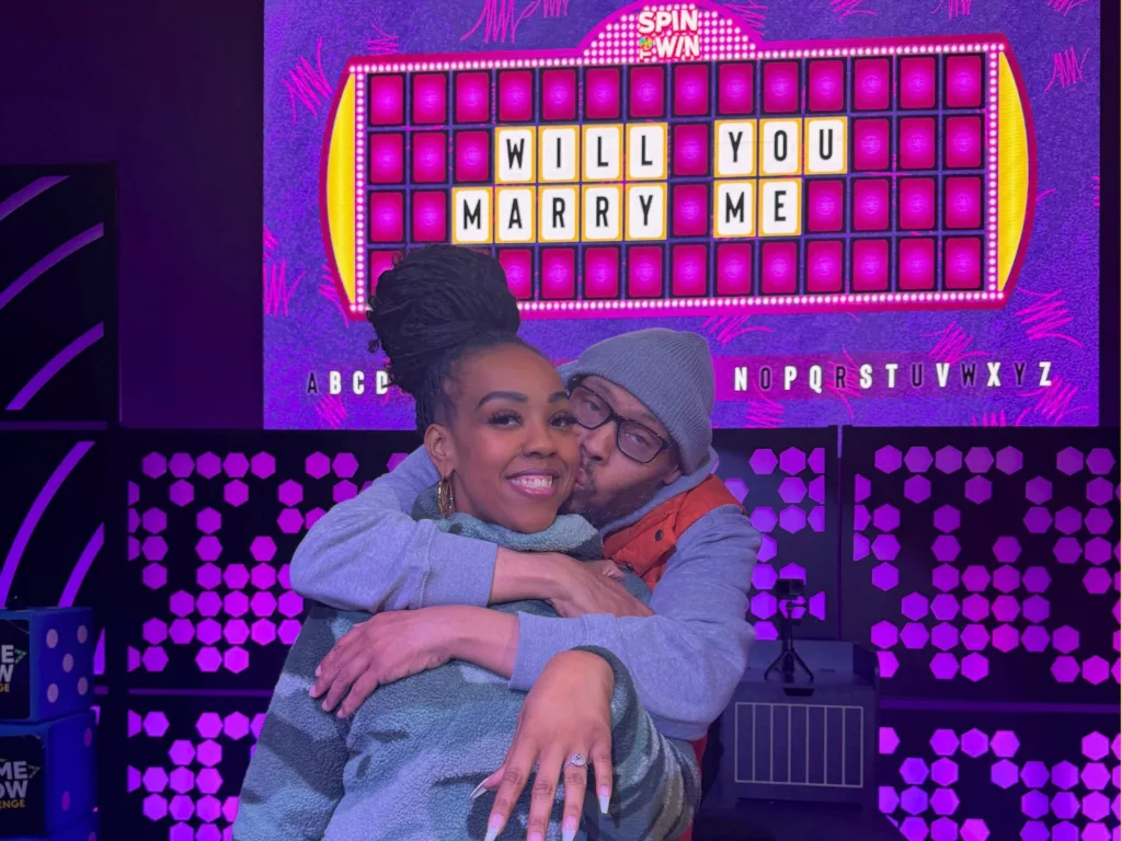 Alicia smiles while Andre hugs her in front of a screen displaying “Will You Marry Me.”