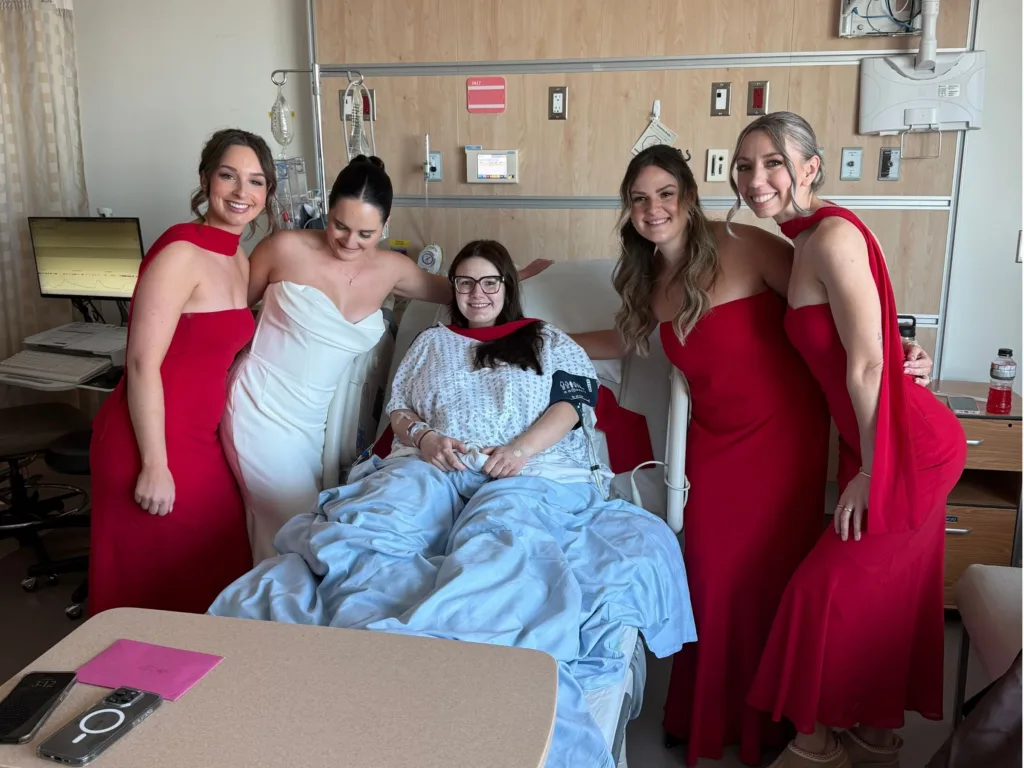 Skye, dressed in white, stands beside Hannah in her hospital bed as bridesmaids in red gather around them smiling.