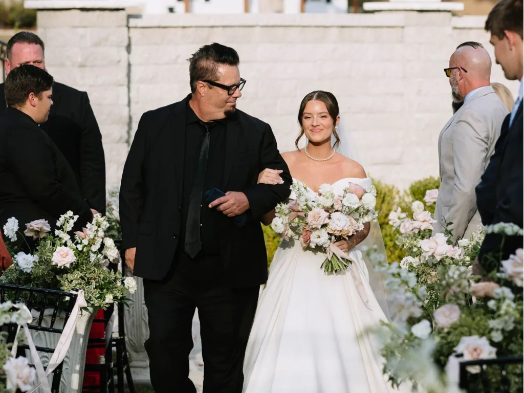 Greg walks Madison down the aisle at her wedding as guests turn to watch and Madison smiles while holding her bouquet.