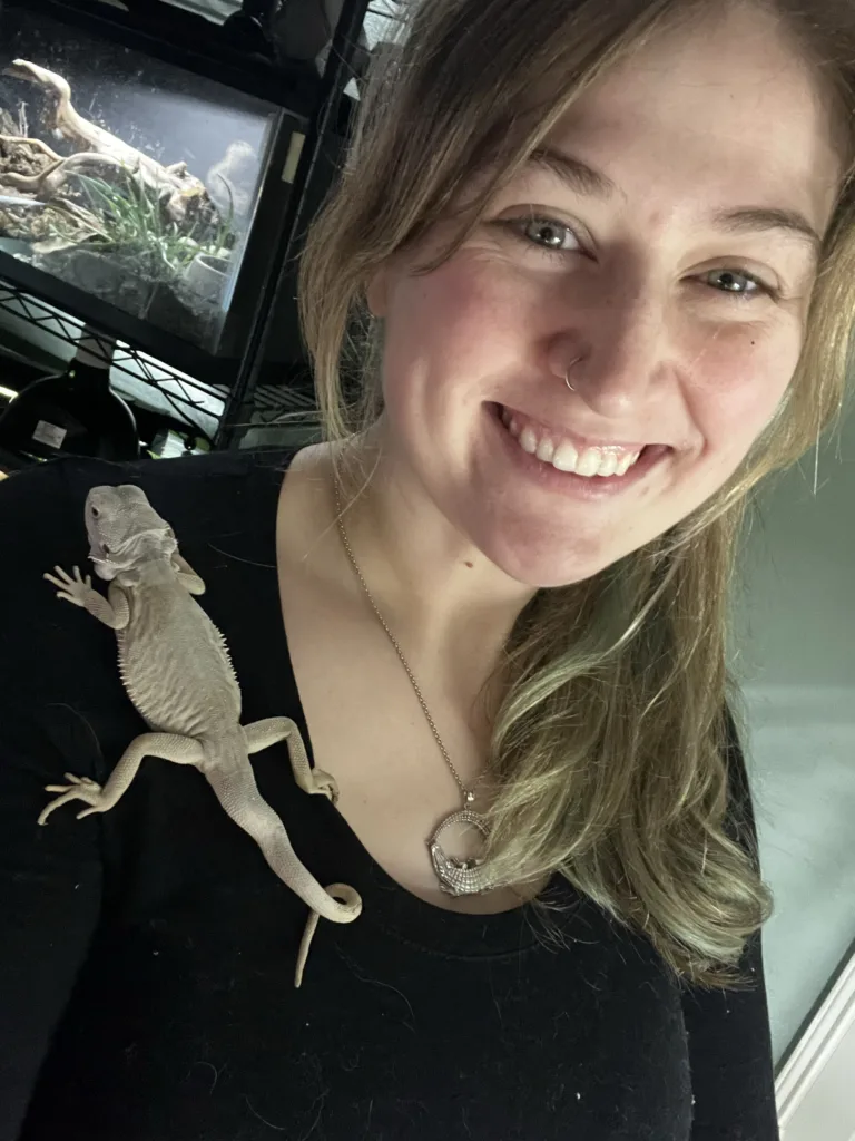 A smiling woman poses for a selfie with a pale bearded dragon resting on her shoulder.