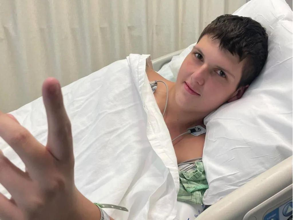Justin lies in a hospital bed and gives a thumbs up during his cancer treatment.