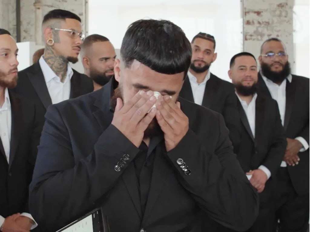Marcos wipes his eyes with a tissue as he stands in a black suit surrounded by wedding guests.