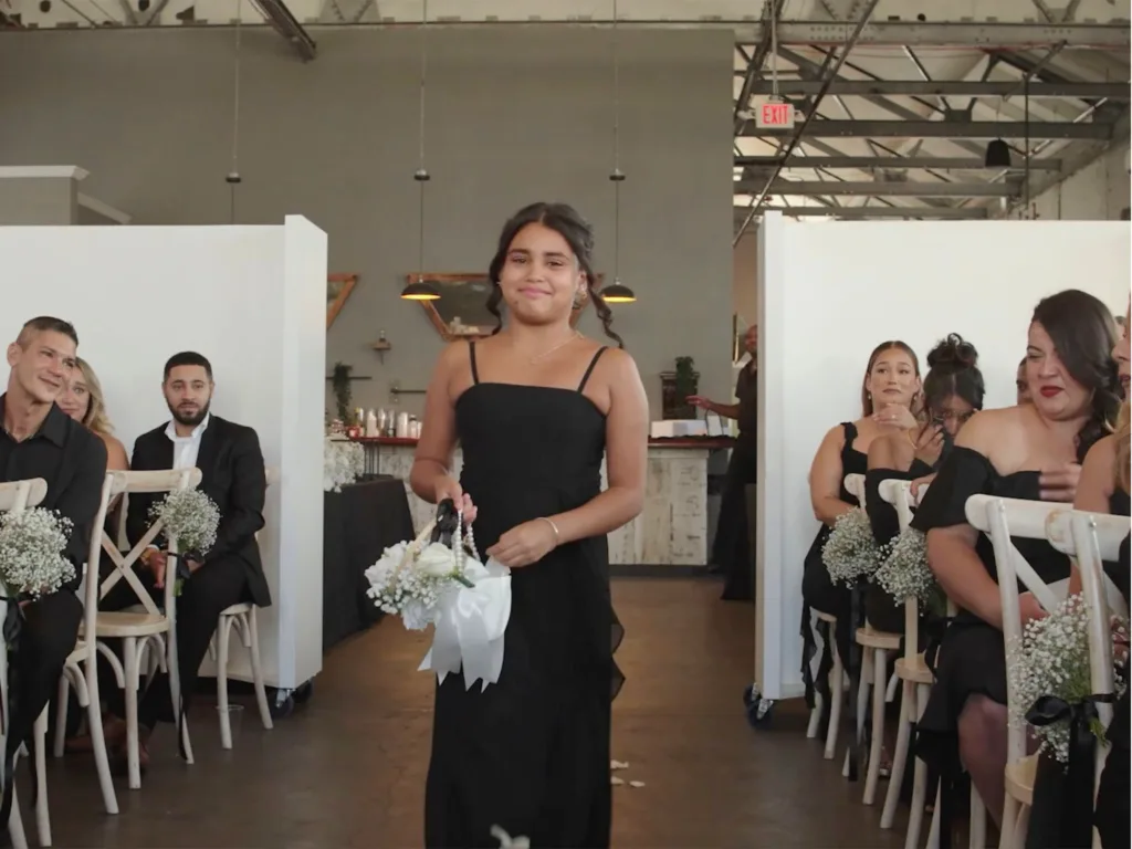 Zoë walks down the aisle in a black dress carrying a white flower basket during Jessica and Marcos’s wedding ceremony.