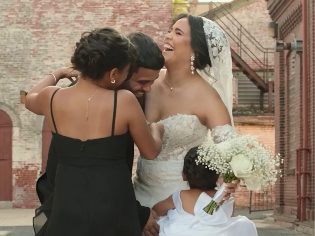 Jessica, Marcos, and Zoë share a joyful group hug outdoors after the wedding ceremony, with a young flower girl holding a bouquet nearby.
