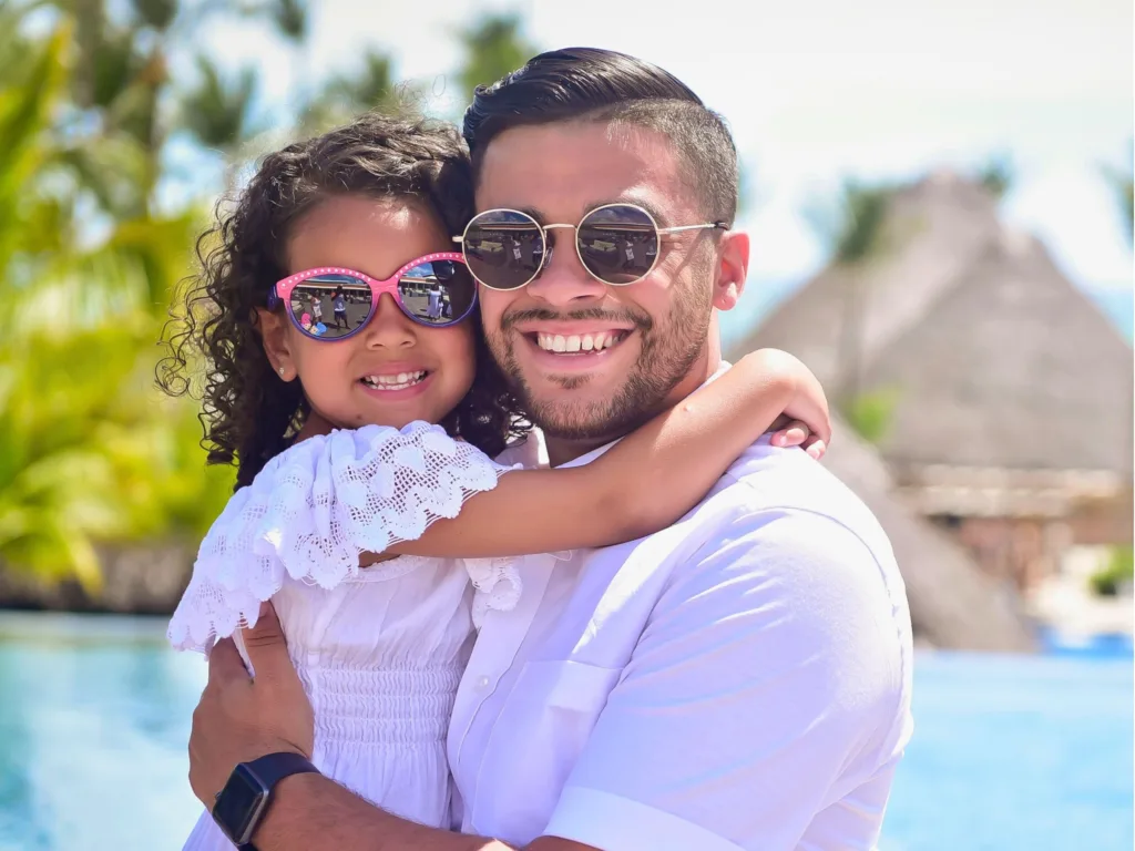 Marcos smiles while holding Zoë outdoors as the pair pose together in matching white outfits and sunglasses.