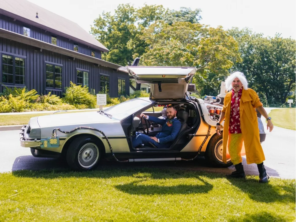 Chris sits inside a Back to the Future DeLorean while a man dressed as Doc Brown stands beside the car