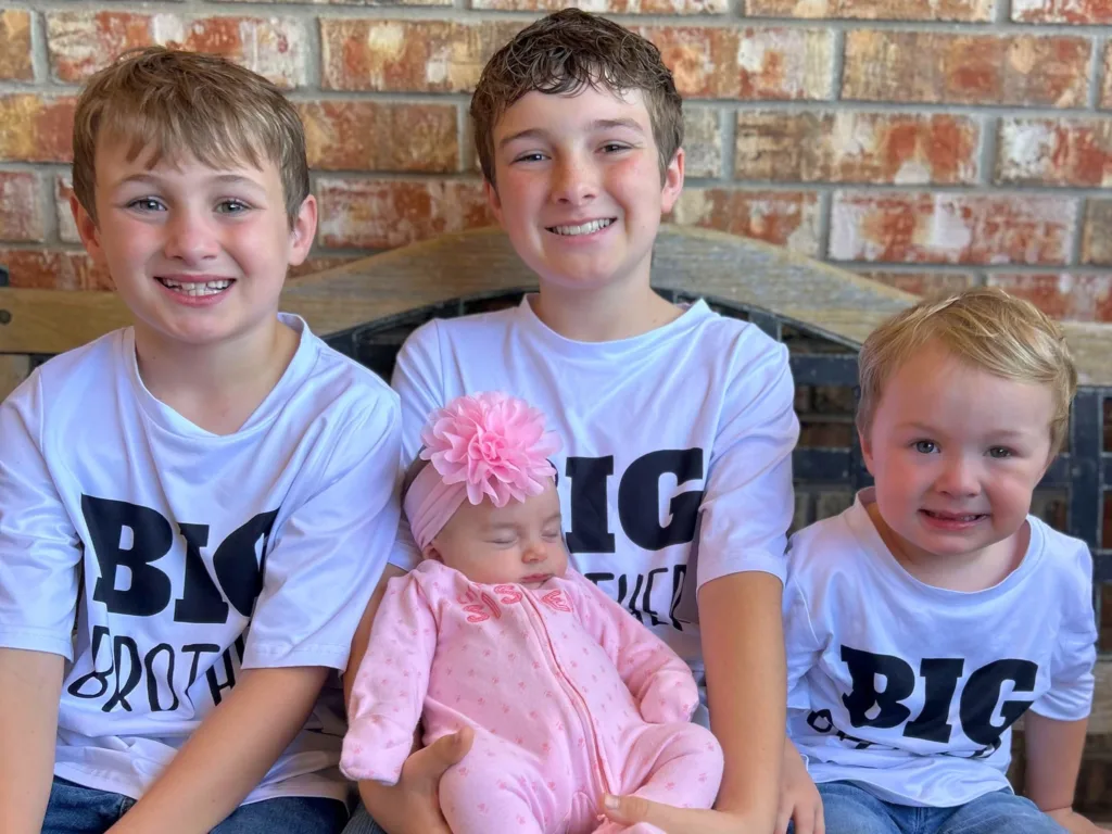 Layken, Liam and Lane sit together holding their newborn sibling dressed in pink while smiling at the camera.