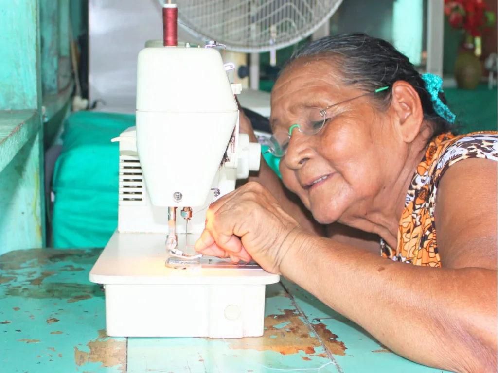 An older woman wearing One Dollar Glasses leans close to a sewing machine to thread a needle.