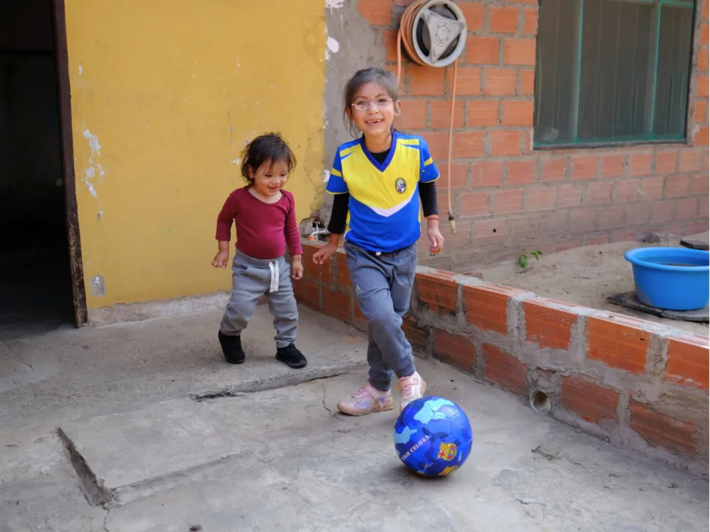 A young girl wearing One Dollar Glasses runs to kick a football in a sunny courtyard while her younger sibling watches.