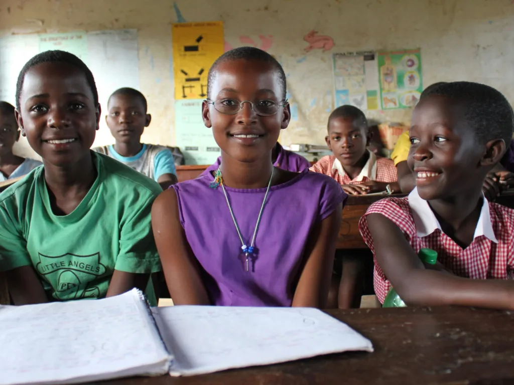 A schoolgirl in a Ugandan classroom wearing One Dollar Glasses smiles between classmates, with a $1 callout pointing to her glasses.