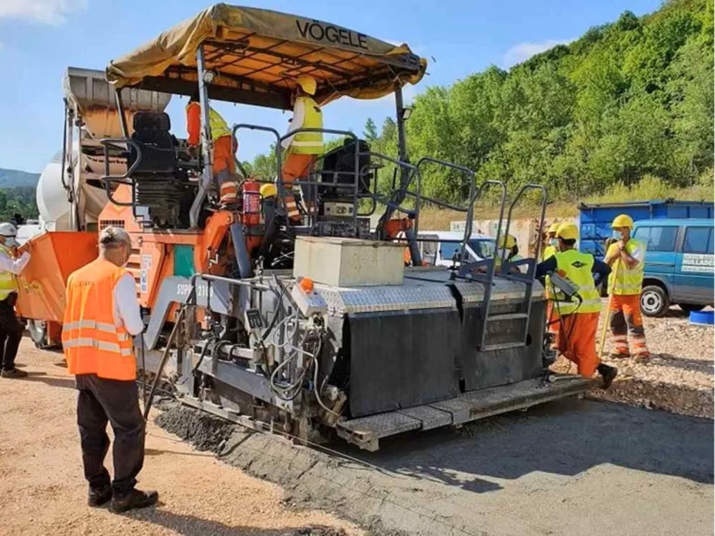 Vögele paving machine laying Waste Light Concrete on a road surface with workers in high-visibility gear.