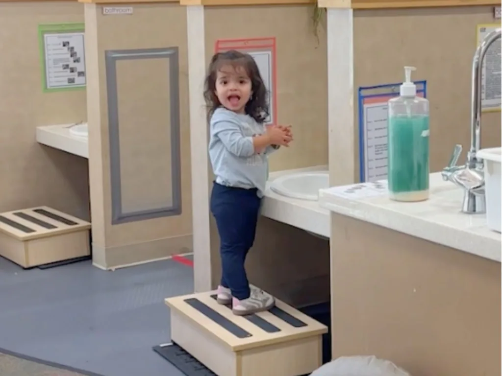 Esme stands on a small step stool by a classroom sink, smiling with surprise as she looks across the room.