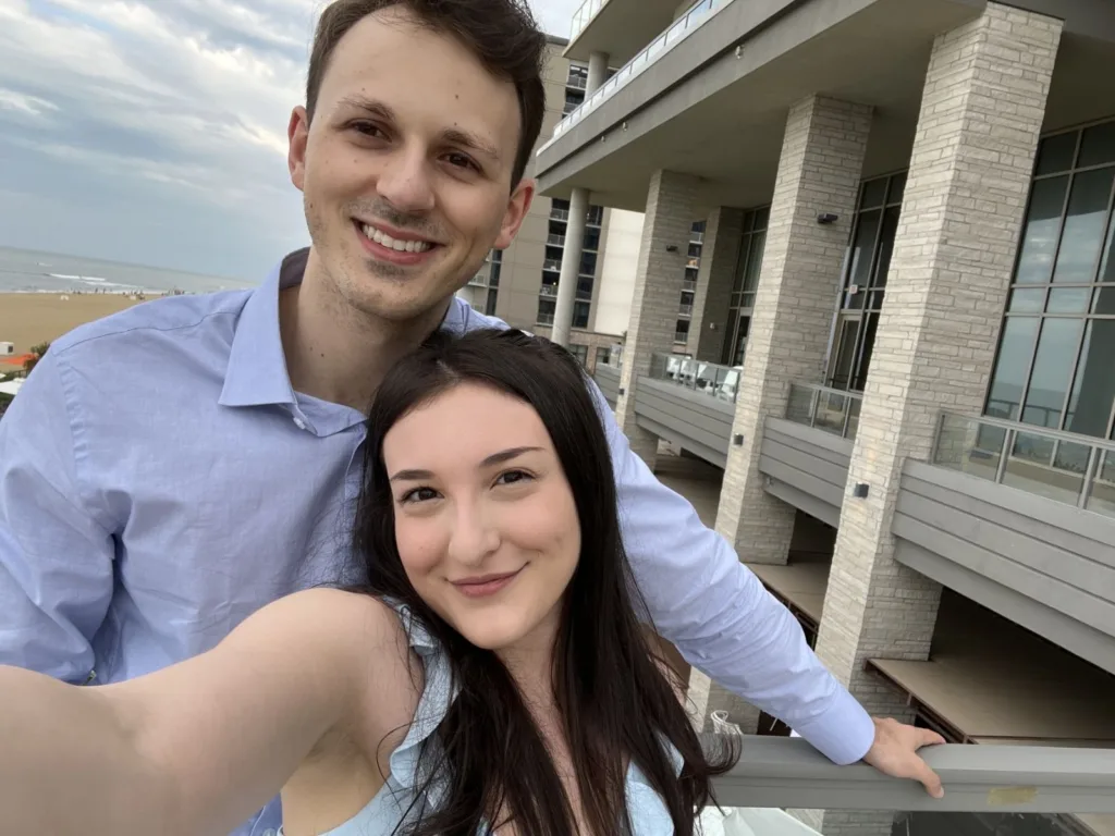 Ila and Matthew smile together in a selfie near the beach.