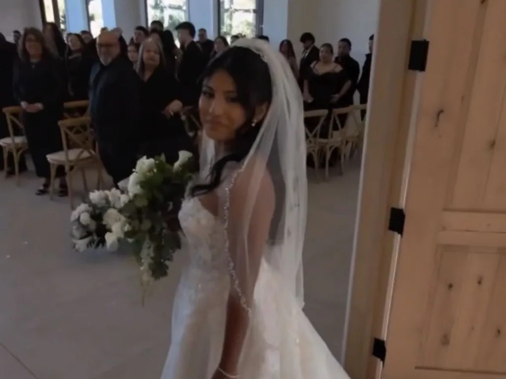 Adrienne stands at the entrance to the wedding ceremony holding a bouquet as guests look on.