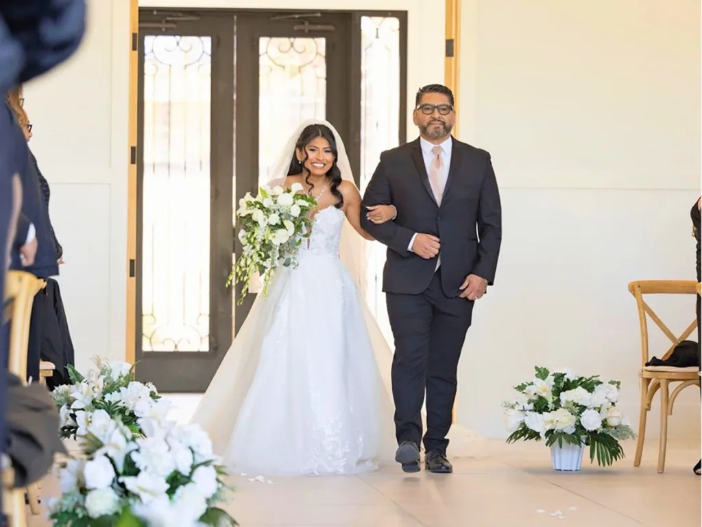 Adrian walks Adrienne down the aisle at her wedding while she smiles and carries a white bouquet.