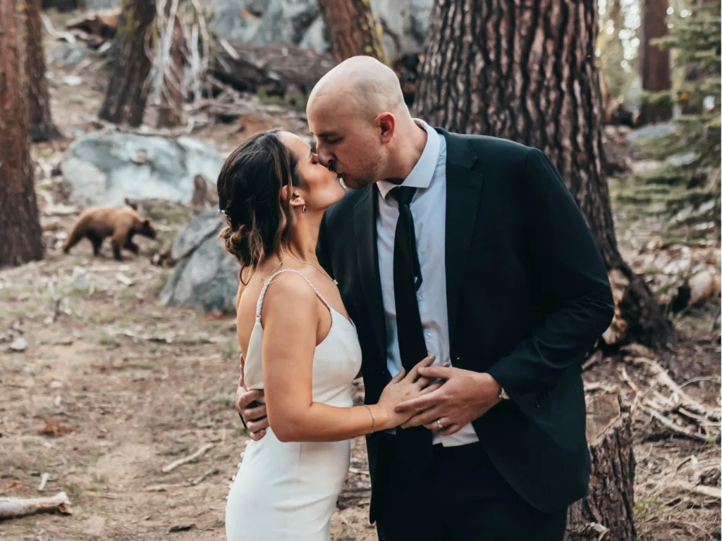 Kelly and Stephen kiss in wedding attire in Yosemite National Park with a wild bear visible behind them.