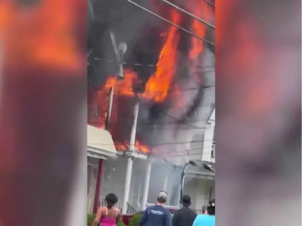 Flames and smoke pour from the upper floor of a house fire in Schenectady as people stand nearby.